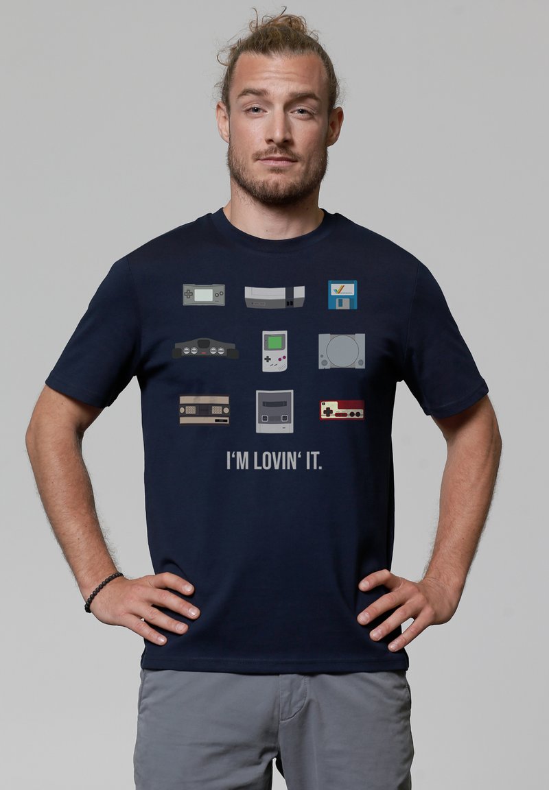 Navy blue t-shirt features pixelated graphics of video game consoles, with the text "I'M LOVIN' IT" below the designs. Cotton material, relaxed fit.