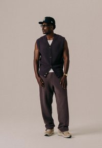 Pleteni ljubičasti prsluk s teksturiranim uzorkom i gumbima, nošen preko bijele košulje, u kombinaciji s opuštenim sivim hlačama i bež tenisicama.