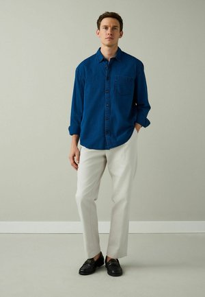 Young man standing indoors wearing a blue button-up shirt, light beige pants, and black loafers against a plain light gray wall.