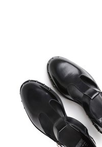 Schwarze Lederschuhe mit T-Bar-Design, Nietenakzenten entlang der Sohle und glatt strukturierter Oberfläche.