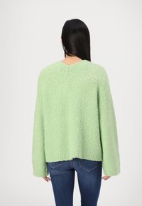 Pull vert clair texturé avec manches longues et larges et encolure en V. L'ourlet inférieur est légèrement raccourci, porté avec un jean foncé.