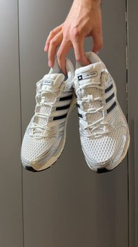 Chaussures de sport blanches avec dessus en mesh, rayures latérales noires et accents réfléchissants. Semelle en caoutchouc crantée, design à lacets, texture lisse.