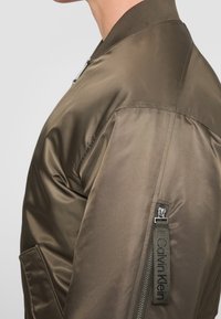 Olijfgroene bomberjack met een gladde satijnen afwerking, geribbelde kraag, zijzak en een ritstrekker met het merk "Calvin Klein" tekst.