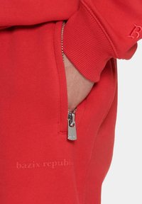 Roter Kapuzenpullover mit gerippten Bündchen, seitlicher Reißverschlusstasche und dezent eingeprägtem "bazix republic"-Text auf dem Stoff. Glatte Textur.