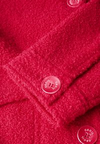 Röd texturerad tyg med en fluffig yta, med två genomskinliga knappar märkta "Cecil" och en sydd placketdetalj.