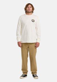 Mann mit schulterlangem Haar, der ein weißes Langarmhemd mit einem runden Logo, khakifarbene Hose und schwarze Sneakers trägt, vor einem weißen Hintergrund steht.