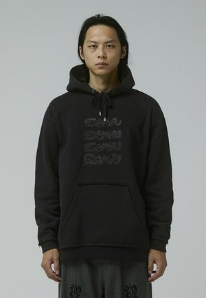 Black hoodie with a kangaroo pocket, featuring tonal text in a wavy pattern across the front. Material appears soft and textured.