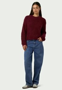 Βουργουνδής αφράτος πουλόβερ με στρογγυλή λαιμόκοψη, συνδυασμένο με χαλαρά μπλε τζιν παντελόνια και γκρι παπούτσια. Το σύνολο έχει άνετη εφαρμογή και casual στυλ.