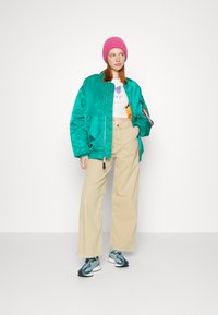 Teal bomberjacka, vit crop top med lila grafik, beige vida byxor, rosa stickad mössa och flerfärgade sportskor.