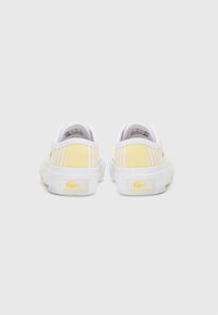 Baskets Lacoste, avec une tige en toile à rayures verticales jaunes, une semelle en caoutchouc blanche et une marque jaune sur le talon.