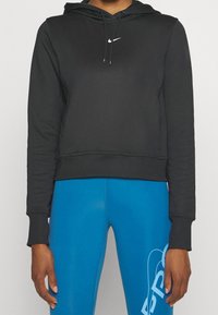 Sudadera negra recortada en material suave con capucha ajustable y logo blanco de Nike. Combinada con leggings azul brillante que llevan un estampado blanco.