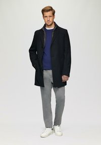Zwarte wollen jas met een gekartelde kraag, over een blauwe trui, gecombineerd met grijze joggingsbroeken en witte sneakers. Soepele textuur, nette pasvorm.