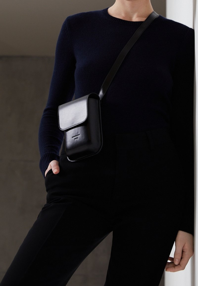 Personne portant un pull et un pantalon noirs, avec un petit sac bandoulière en cuir noir à rabat.