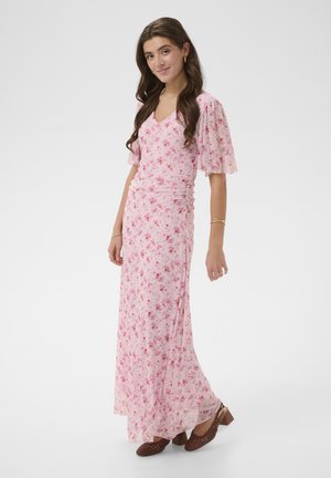 Jonge vrouw met lang haar, gekleed in een roze maxi-jurk met bloemenprint en een V-hals, en bruine gevlochten schoenen met blokhak, glimlachend en staand.