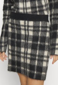 Ullklänning med ett svart och vitt rutmönster, med långa ärmar och en figursydd midja, mjuk textur med en något fluffig yta.