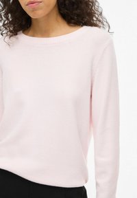 Femme portant un pull rose clair à col rond et un pantalon noir, montrée de la poitrine jusqu'aux hanches sur un fond blanc.