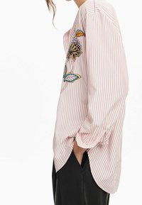 Langärmliges gestreiftes Hemd in Hellrosa mit besticktem Blumenmuster in verschiedenen Farben, mit lockerem Schnitt und seitlichem Schlitzdetail.