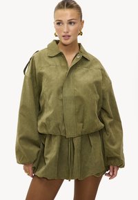 Femme portant une veste oversize vert olive avec un col et une fermeture éclair, associée à un short ample assorti, des boucles d'oreilles créoles en or et des bagues.