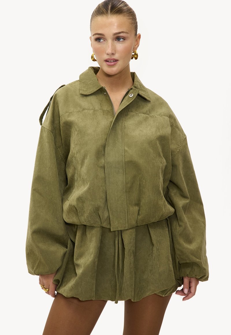 Femme portant une veste oversize vert olive avec un col et une fermeture éclair, associée à un short ample assorti, des boucles d'oreilles créoles en or et des bagues.