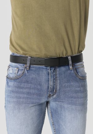 Hombre con jeans azul claro y camisa verde oliva con un cinturón de cuero negro abrochado alrededor de la cintura.