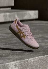 Sapatilha desportiva em camurça rosa com detalhes castanhos, bico perfurado, atacadores lisos e sola de borracha, posicionada acima de uma superfície de betão.