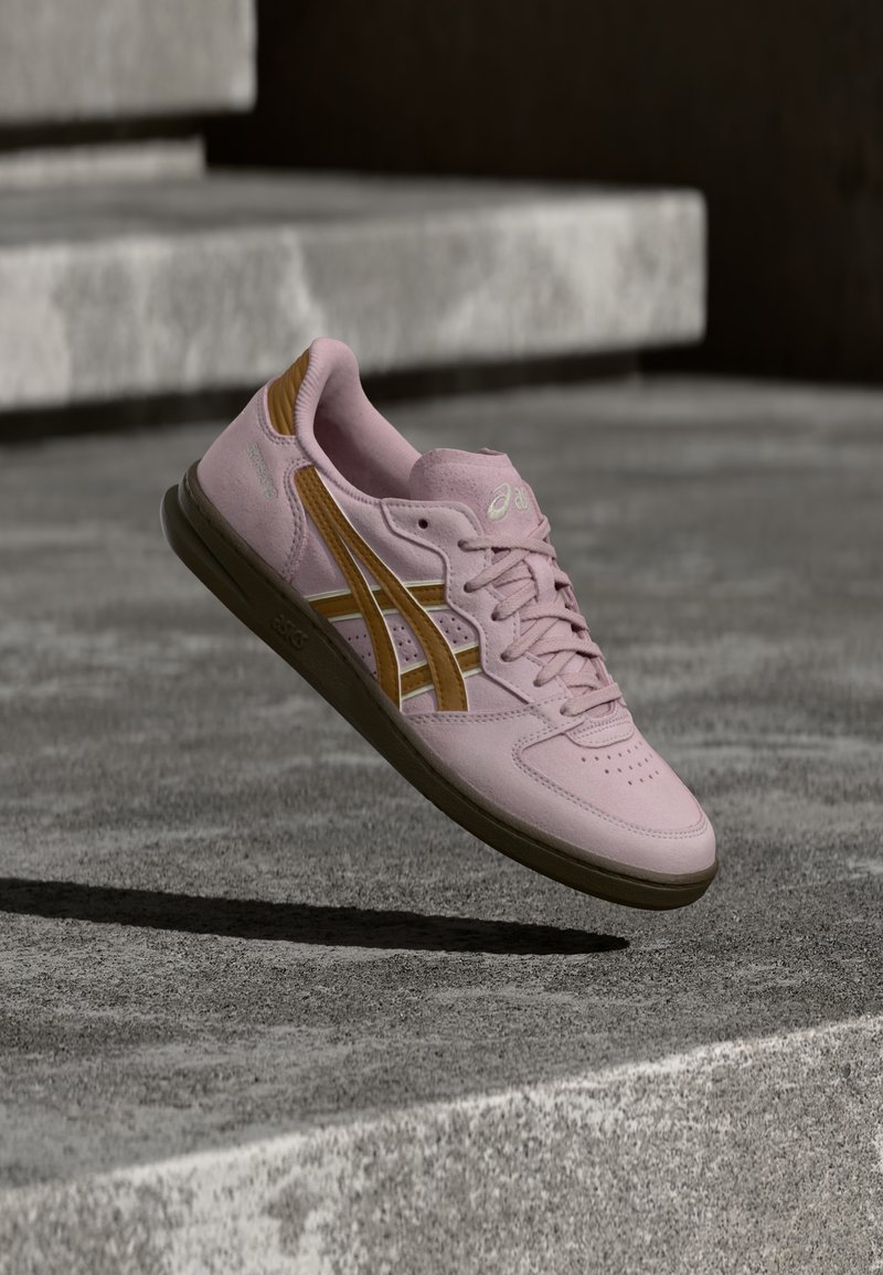 Sapatilha desportiva em camurça rosa com detalhes castanhos, bico perfurado, atacadores lisos e sola de borracha, posicionada acima de uma superfície de betão.