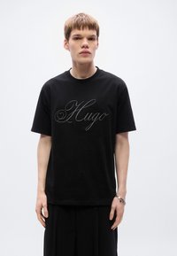 Μαύρο βαμβακερό t-shirt με κοντά μανίκια, με μεγάλο γκρι λογότυπο "Hugo" σε γραφικό σενάριο στο μπροστινό μέρος. Λαιμός τύπου crew, άνετη εφαρμογή.