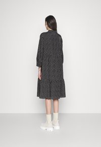 Eine Frau, die mit dem Rücken zur Kamera steht, trägt ein schwarzes gemustertes Midikleid mit langen Ärmeln und weiße, klobige Sockenschuhe vor einem einfarbigen Hintergrund.