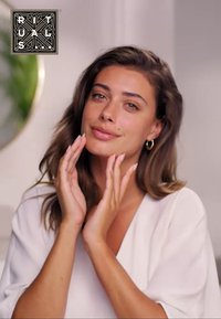 Modèle avec de longs cheveux bruns ondulés, portant un haut blanc. Elle sourit tout en touchant son visage, mettant en valeur une texture de peau naturelle.