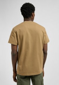 T-shirt di cotone color cachi con maniche corte e collo tondo. Presenta un taglio dritto e una texture liscia, indossato con pantaloni cargo verdi.
