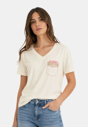 Vrouw met lang bruin haar die een witte V-hals T-shirt met rode tekst en een blauwe spijkerbroek draagt, hand in de zak, neutrale achtergrond.