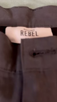 Črn material z gladko teksturo, s etiketo, na kateri piše "REBEL", pritrjeno na žepu z majhno luknjico za gumb.