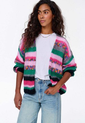 Kolorowy, dziergany kardigan z szerokimi rękawami, ozdobiony wyraźnymi paskami w kolorach różowym, zielonym, szarym i czarnym. Nosi się go na białą koszulkę i dżinsy.