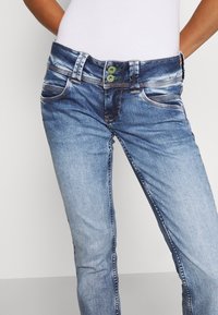 Blå denimjeans med en blekt tvätt, rak ben, två framfickor och en midja med två gröna knappar.