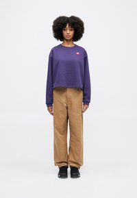Purple cropped sweatshirt with a small red logo, paired with loose beige pants and black shoes. Model stands against a light background.