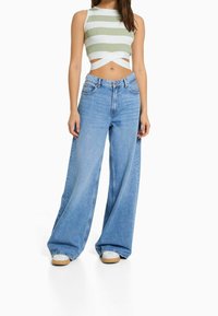 Nainen, jolla on päällä vihreä-valkoraidallinen lyhyt toppi, jossa on ristikkäiset hihnat, sekä korkeavyötäröiset leveälahkeiset siniset farkut ja valkoiset tennarit.