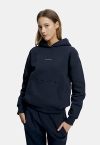 Marineblå hettegenser med lommer foran, ribbestrikkede mansjetter og nederkant. Har en hvit logo "CASPARA" på brystet. Myk tekstur.