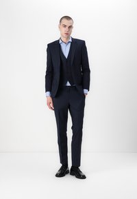 Marineblauw drie-delig pak van soepel materiaal, met een getailleerde blazer, een vest met knopen en slanke broeken, gecombineerd met zwarte schoenen.