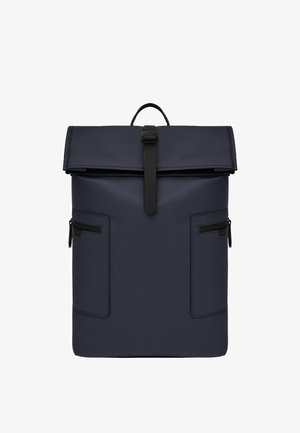 Tummansininen rullattava reppu mustalla solkikiinnityksellä, kahdella sivutaskulla, joissa vetoketjut, ja yläosassa kantokahva valkoisella taustalla.