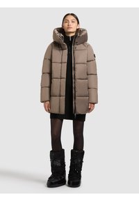 Padded brown coat with a hood, featuring a zip closure and quilted design. Worn over a black sweater dress with knee-high black boots.