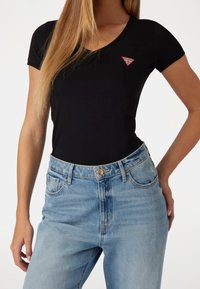Zwarte korte mouwen t-shirt met een V-hals en een rood driehoekig logo op de linkerborst, gecombineerd met lichtblauwe hooggetailleerde spijkerbroek.