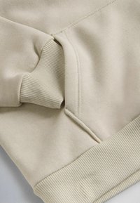 Manica di felpa beige con polsino a costine e orlo a costine visibili, che mostrano i dettagli delle cuciture e la morbidezza del tessuto.