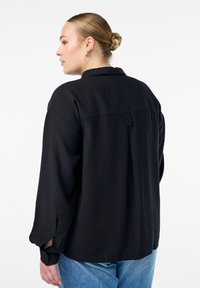 Crna košulja s dugim rukavima i opuštenim krojem, sa detaljima nabora na leđima. Izrađena od lagane tkanine, s preklopljenim manžetama.