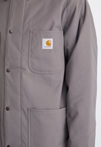 Grå vandtæt jakke med en brystlomme med et hvidt label og orange logo. Trykknaplukning og tekstureret stof.