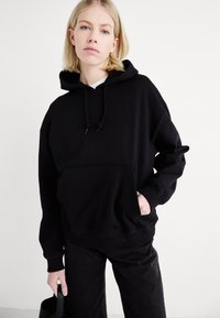 Felpa nera oversize realizzata in tessuto morbido, con tasca frontale, cappuccio con coulisse e polsini a costine. Abbinata a pantaloni neri.