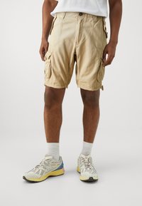 Beige cargo shorts with multiple side pockets, a relaxed fit, stitched detailing, and a hem that stops just above the knee.