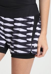 Svarta träningsshorts med ett lagerdesign som har ett vitt zickzackmönster och små prickar. De har en svart midja och innertyg.