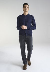 Granatowy kardigan z kołnierzem i zapięciem na guziki, w zestawieniu z ciemnoszarymi dżinsami i brązowymi mokasynami, stojący na neutralnym tle.