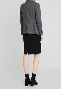 Blazer sartoriale in bianco e nero a fantasia houndstooth con silhouette aderente, abbinato a una gonna a tubino nera con spacco posteriore e stivaletti.