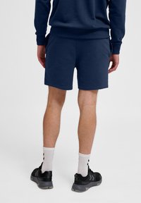 Pantalones cortos de algodón azul marino con cintura elástica, bolsillos laterales y una textura suave. El modelo lleva zapatillas negras y calcetines blancos.